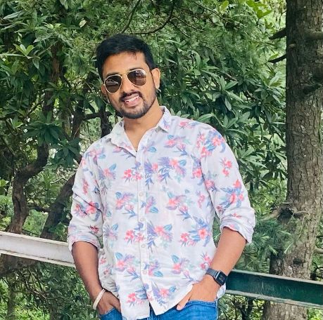 Aditya Raj Singh with short dark hair and a beard, wearing a white shirt and dark suit jacket, poses against a plain dark background.