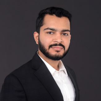 Aditya Raj Singh with short dark hair and a beard, wearing a white shirt and dark suit jacket, poses against a plain dark background.